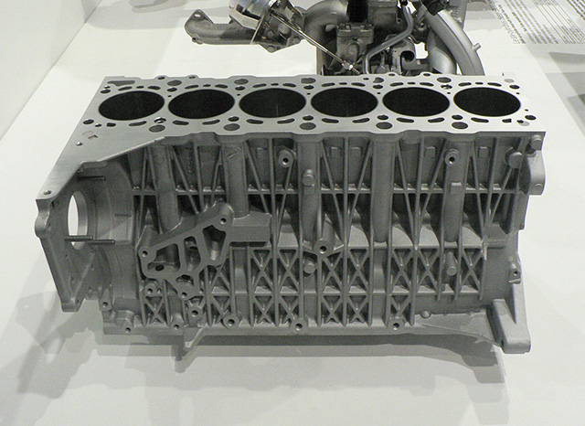 Sobre una superficie blanca se muestra un bloque de motor met&aacute;lico de seis cilindros. La estructura, probablemente producida por un proveedor de fundici&oacute;n a presi&oacute;n en China, presenta patrones geom&eacute;tricos intrincados y varios puntos de montaje, lo que muestra su ingenier&iacute;a detallada. Al fondo se ve parcialmente un complejo conjunto de tuber&iacute;as