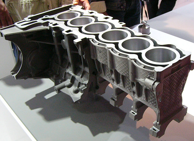Un primer plano de un bloque de motor seccionado, que muestra la intrincada estructura interna y los orificios de los cilindros. El componente met&aacute;lico est&aacute; expuesto sobre una mesa, mientras varias personas lo observan al fondo. La imagen resalta los detalles de ingenier&iacute;a y dise&ntilde;o del motor, cortes&iacute;a de una empresa de fundici&oacute;n a presi&oacute;n de aluminio