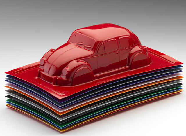 Un modelo rojo brillante de un autom&oacute;vil cl&aacute;sico, que se asemeja a un Volkswagen Beetle, est&aacute; colocado encima de una ordenada pila de coloridas l&aacute;minas de pl&aacute;stico rectangulares. El autom&oacute;vil y las l&aacute;minas exhiben una superficie brillante y reflectante, que muestra el moldeado al vac&iacute;o de pl&aacute;stico personalizado con calidad de clase mundial contra un simple degradado de blanco a gris